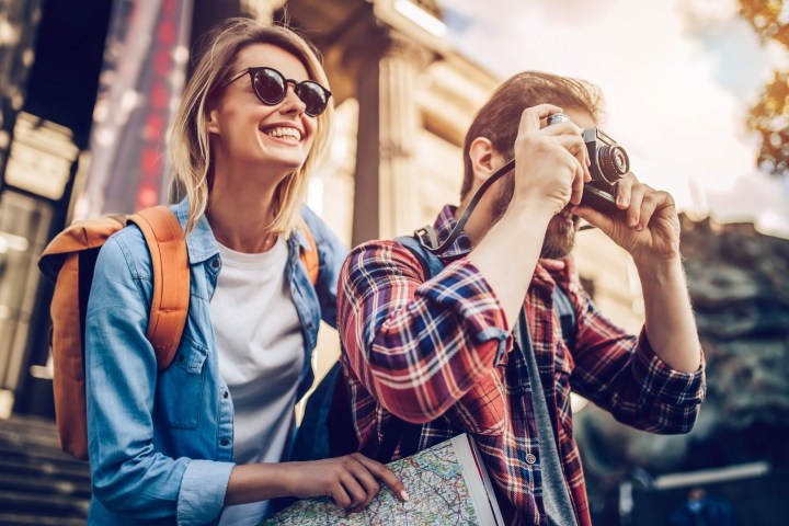 A smiling couple outdoors, one with a camera, the other holding a map, both wearing backpacks.
