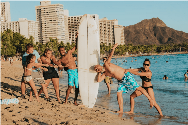 blog2 People playing with a surfboard on a beach, with cityscape and mountain in the background.