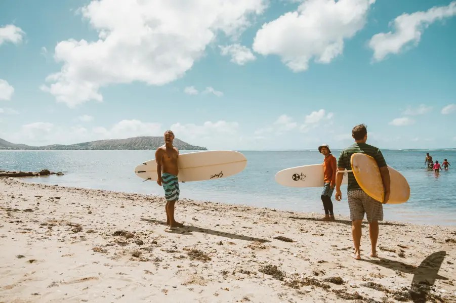 Three people with surfboards on a sandy beach under a blue sky.