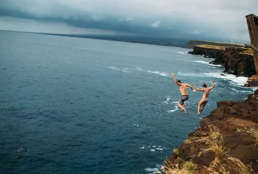 Two people cliff jumping into the ocean under cloudy skies.