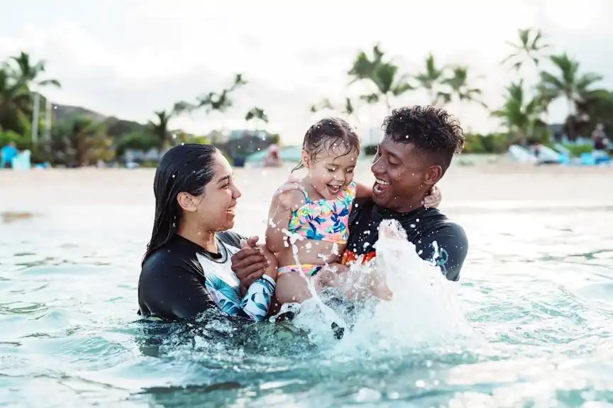 Family of three playing in ocean water near a beach with palm trees.