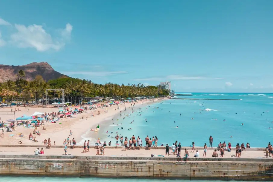 Crowded beach with people swimming and sunbathing, mountain in background, clear blue sky.