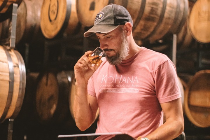 A man in a cap and pink shirt sniffs a drink in a barrel room, holding a clipboard.