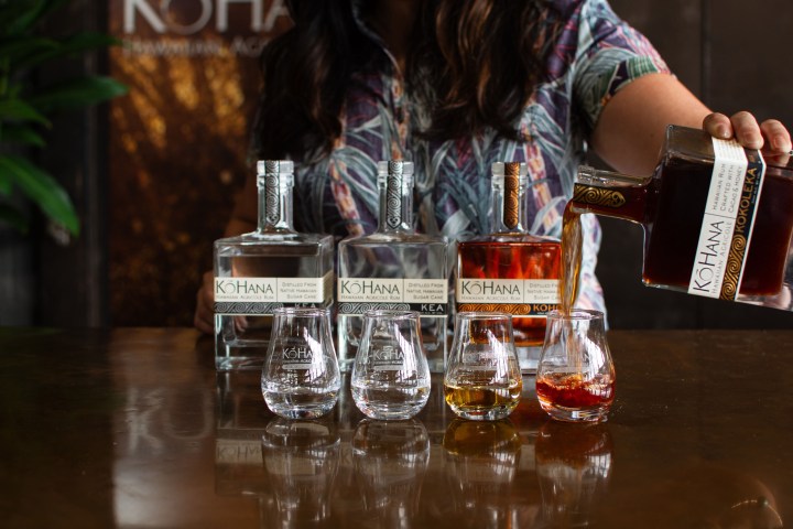 Person pouring dark rum into a glass, with Kohana rum bottles on a wooden table.