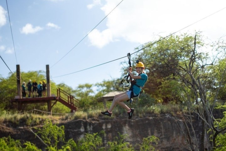 Person ziplining through a forested area on a sunny day, wearing a helmet and harness.
