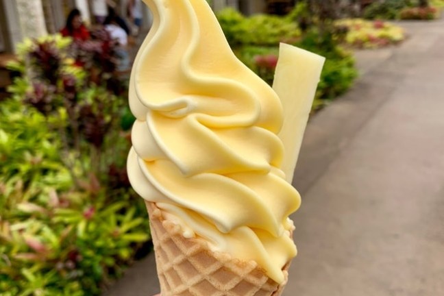 Hand holding a waffle cone with yellow soft serve ice cream and a pineapple slice.