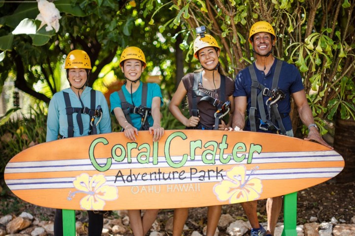 Four smiling people wearing helmets stand behind a 'Coral Crater Adventure Park' sign outdoors.