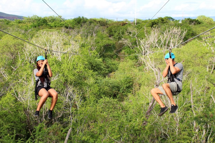 Two people zip-lining over lush green forest wearing helmets and harnesses.