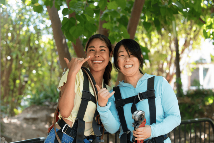 Two women in harnesses smiling under trees, giving a thumbs-up and shaka sign.