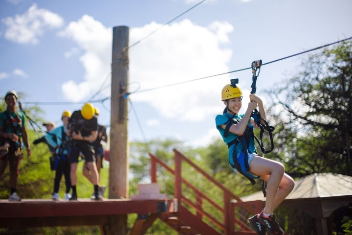 Person ziplining with helmet and harness, others waiting on platform, sunny day.