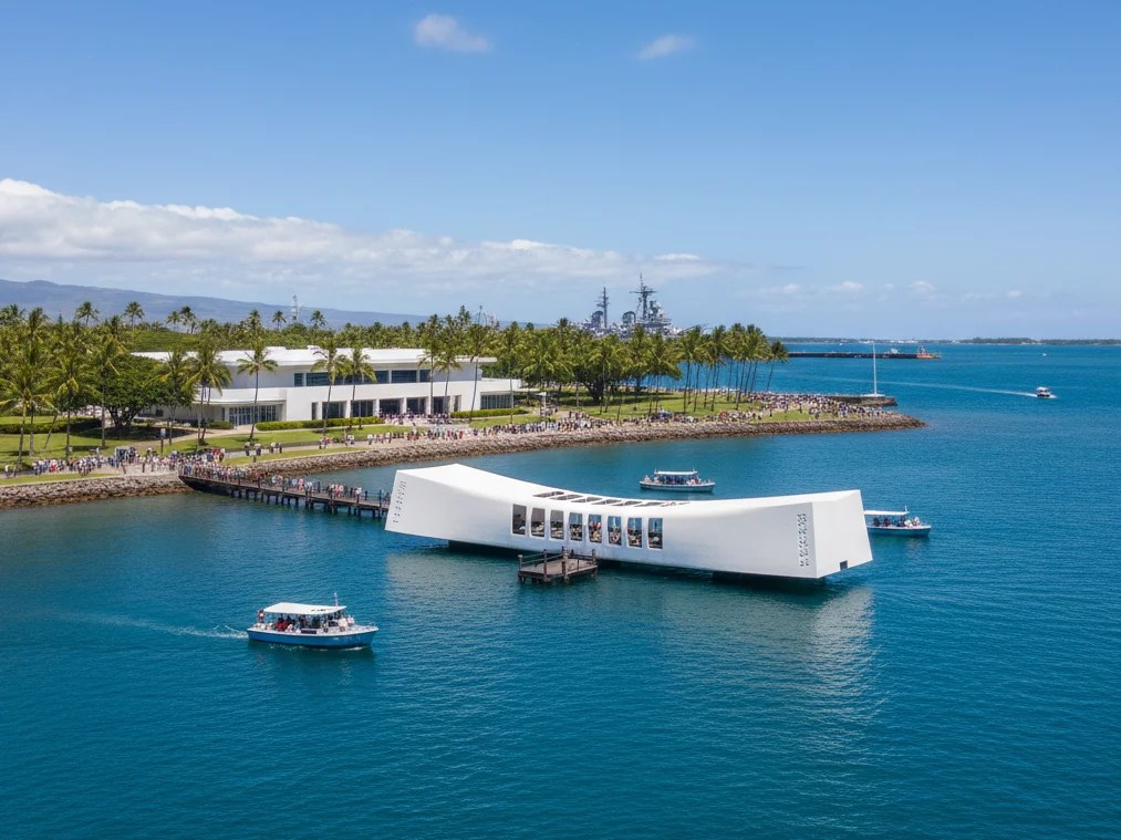 Arial view of white memorial structure on water with boats and palm trees in the background under blue sky.