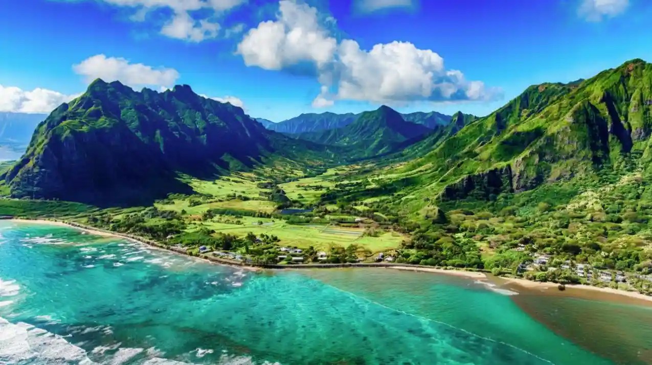 Lush green mountains and turquoise ocean under a bright blue sky with clouds.